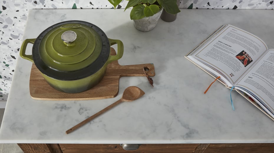 Marble countertop with green dutch oven, wooden cutting board, wooden spoon and an open cookbook